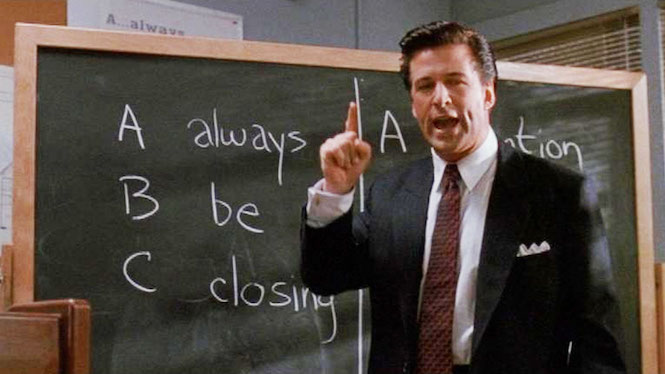 A middle-aged man dressed in a white dress shirt, tie, and blue suit, standing in front of a blackboard on which is written 'Always Be Closing', speaking while his right-hand index figure is raised