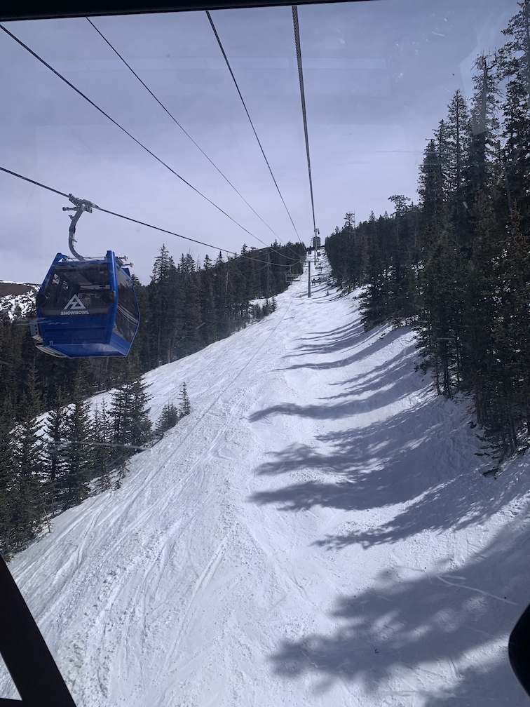 View from inside a gondola car looking up a steep snow-covered mountainside towards the top of the lift, with another gondola car on the left