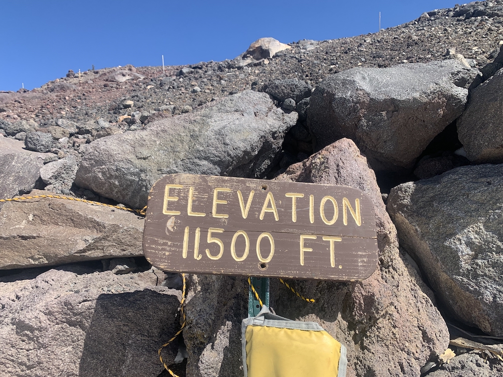 Wooden sign which reads Elevation 11,500 feet, with a rocky mountainside and clear blue sky behind it