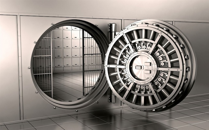 A black and white photo of an open circular vault door that is large enough for a person to walk through, behind which is a room with a wall of smaller closed doors