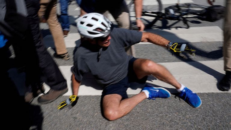 A bicyclist, wearing a bike helmet, shorts, and gloves, starting to get up from the pavement after a fall