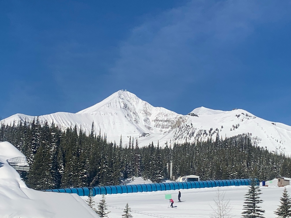 A large, snow-capped mountain against a nearly clear, blue sky, with thick pine trees at lower elevation, and in the foreground two people skiing across a gentle slope in front of a blue windbreak