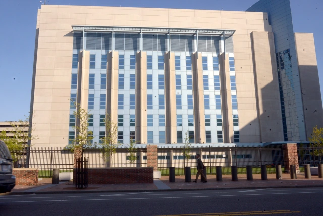 A modern industrial building of approximately seven stories with a beige concrete facade and twelve colums of small blue-tinted windows, behind an iron fence, a sidewalk, and a city street