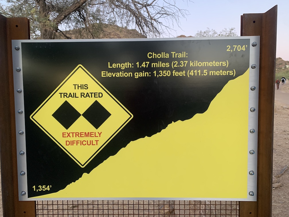 A yellow and black metal sign, with a large yellow diamond containing two smaller black diamonds and the words 'extremely difficult' in all caps, and a jagged yellow area that rises to the upper right edge, along with labeling discussing the elevation gain and length of the Cholla Trail