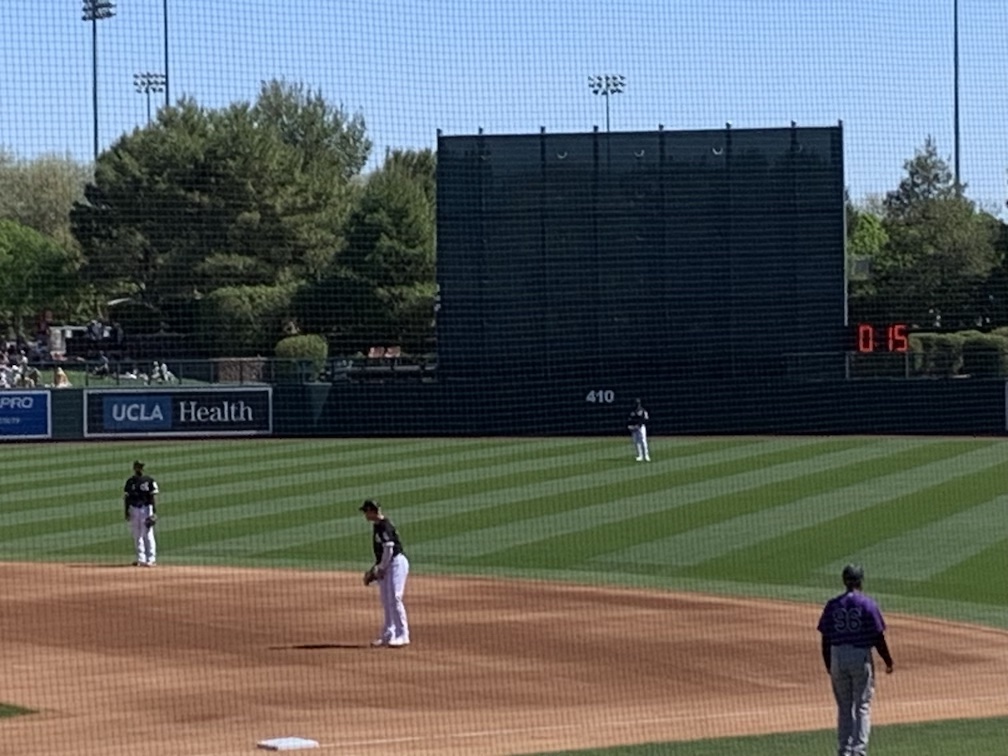 A view of first base and centerfield of a baseball field, with three defenders in the foreground, and beyond the centerfield fence a digital clock showing zero minutes and fifteen seconds of time