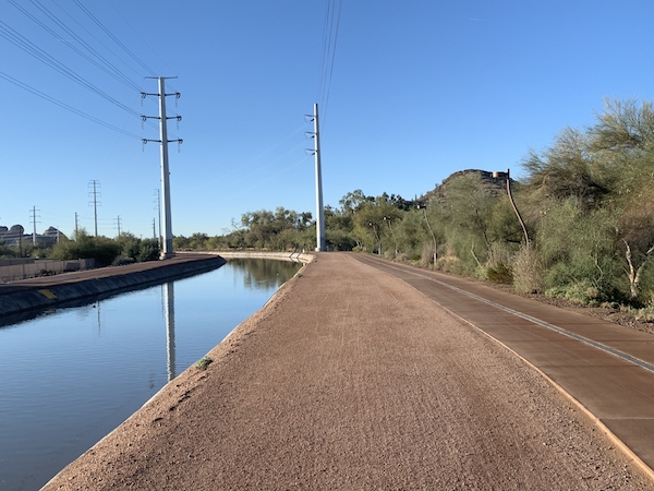 Calm water in a canal that curves to the left, next to smooth dirt and a paved bike path in a desert landscape