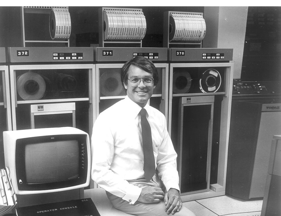 A black-and-white photo of a man wearing glasses, a dress shirt, tie, and slacks, smiling at the camera while seated next to a computer terminal on a table in a computer room, with large computing machines in cabinets in the background