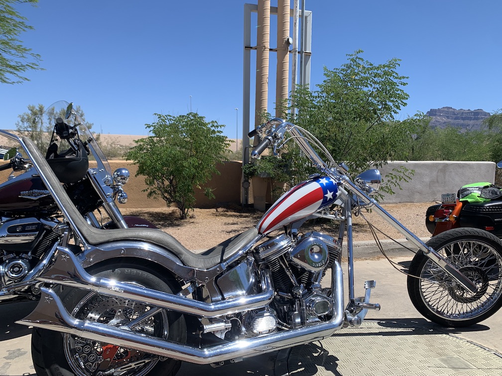 A customized chopper motorcycle, with a gas tank painted to resemble the U.S. flag, and lots of chrome on the engine, frame, forks, and handle bars, with a tall chrome seat and sissy bar covered in dark padding
