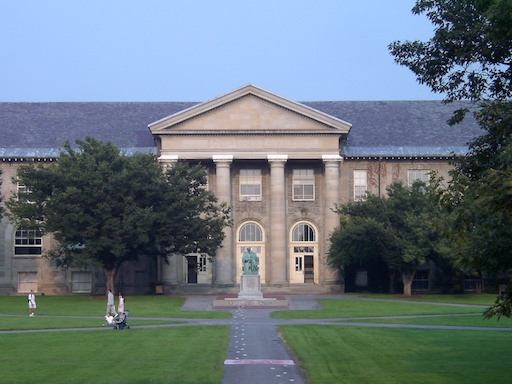 An expansive green lawn with paved paths and mature trees, with a large two story academic building in the background, with a statue in front of it