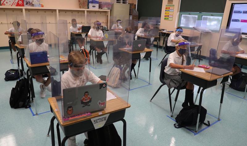 Schoolkids in a classroom sitting behind plexiglass at their socially distanced desks, while wearing masks and face shields