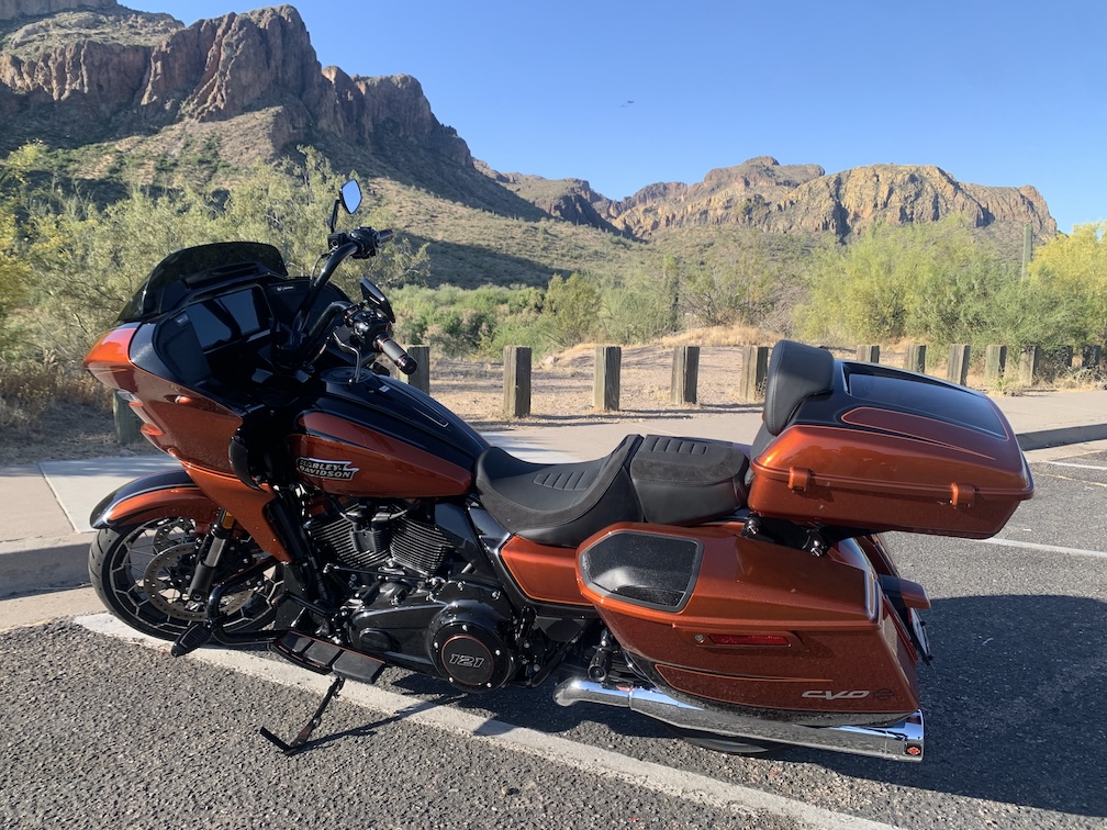 A large orange and black motorcycle, viewed from the left side, parked on an asphalt parking lot with mountains and desert vegetation in the background