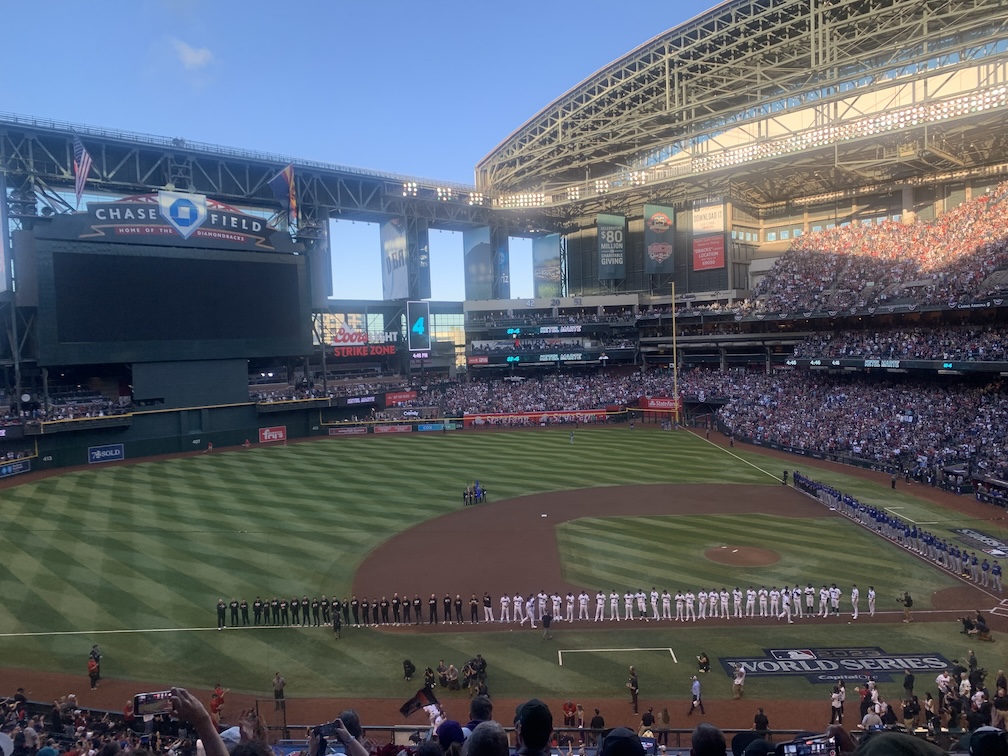 A view of a baseball field taken from the third base stands, with players and staff for both teams lined up along the baselines from the outfield to home plate, beyond which are the stands of the stadium which are packed with fans, and above which are lights and a retracted metal roof