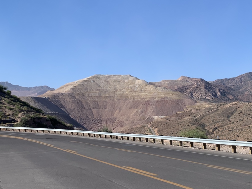In the foreground a smooth asphalt road with a guard rail, in the background some mountains, several of which have had their natural surfaces removed and turned into eroding mounds of rock and dirt traversed by earthen ramps