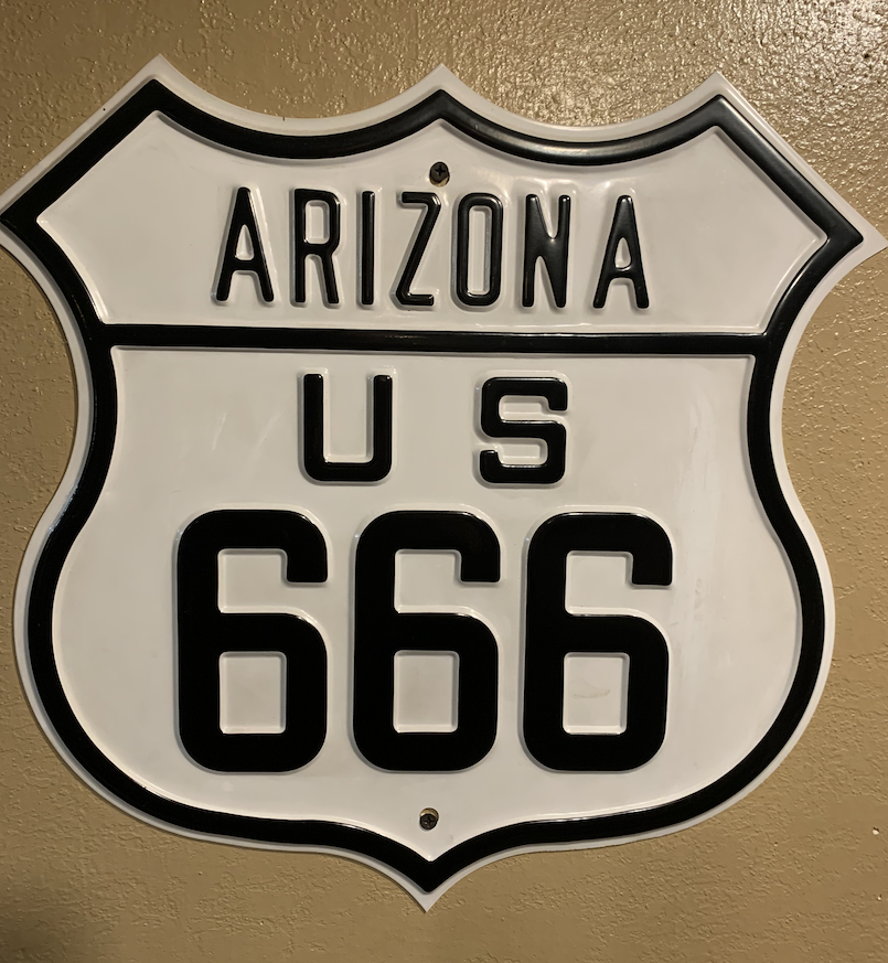 A white badge-shaped highway sign with black lettering and a black border, hanging on a beige wall, for US 666 in Arizona