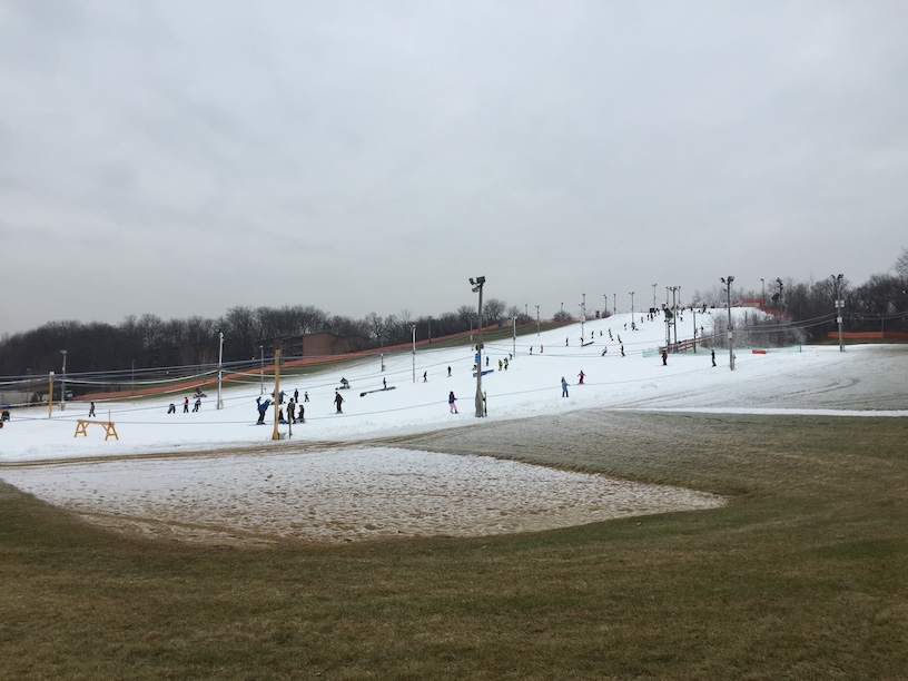 In the background a gentle hill patially covered in snow, with many equipment poles and ropes on the hill along with a few dozen people, and in the foreground some grass that appears to be dormant for the winter