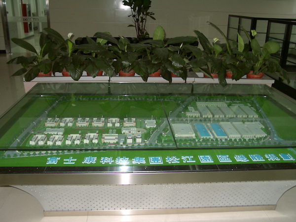An architect's model, with a transparent protective covering, showing a campus of buildings represented as small cardboard boxes, with taller buildings on the left and larger-footprint, shorter buildings on the right