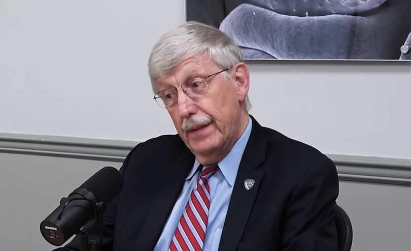 Francis Collins in a suit and tie, seated in front of a microphone, with an expression on his face which indicates he'd prefer to be discussing something else