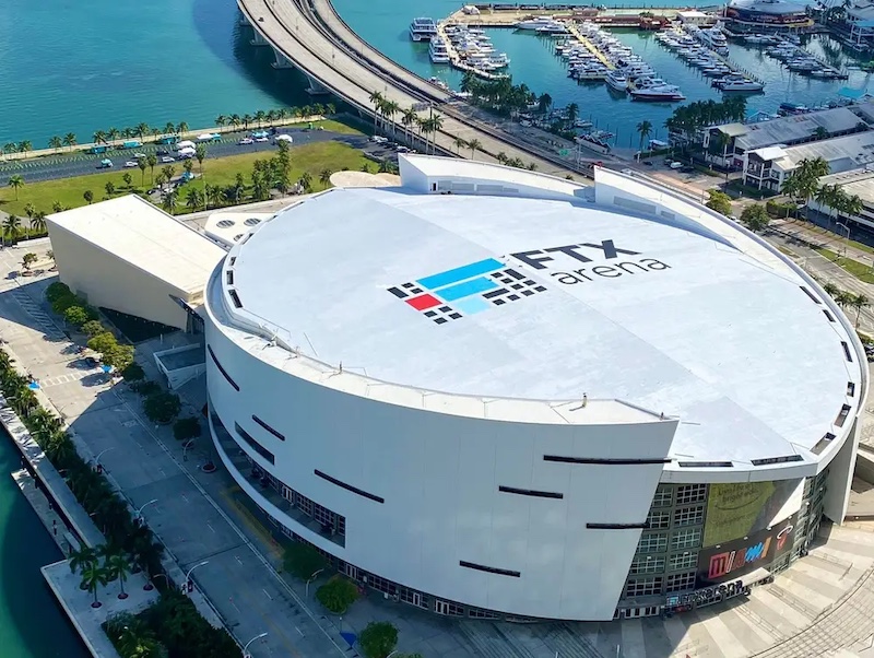 A view from above of a large, oval-shaped, mostly white building, with glass covering one portion of the exterior wall, and a stylized letter 'F' logo along with the words 'FTX arena' painted on the roof, with a parking lot, highway, bridge, water, and marina in the background