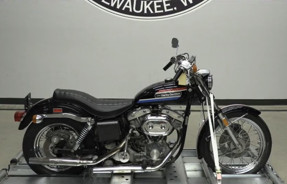 A black 1974 Harley-Davidson FXE 1200 cruiser motorcycle, viewed from the right side while held upright in a metal stand, inside a room