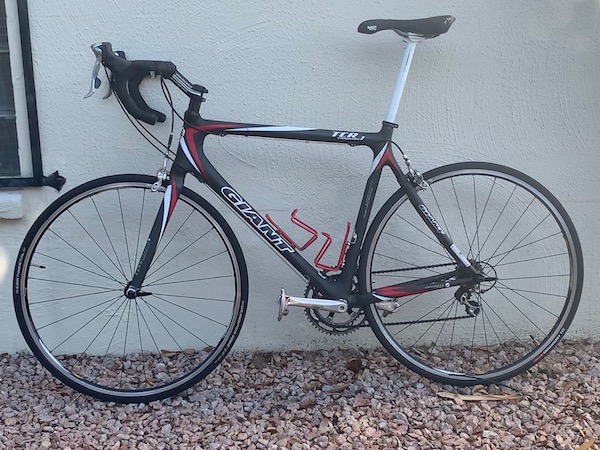 A black-framed racing-style bicycle, with drop handle bars and a tall white seat post, viewed from the left side while it is positioned on a rock bed leaning against a stucco wall