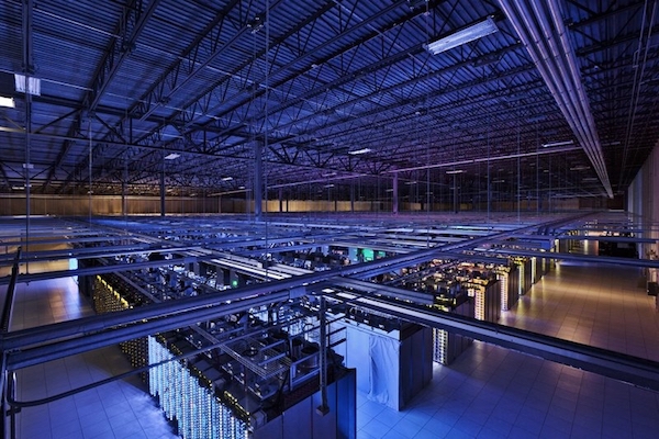 A football-field sized darkened data center, with many rows of racks of computer equipment, and pipes running overhead, and no human being present