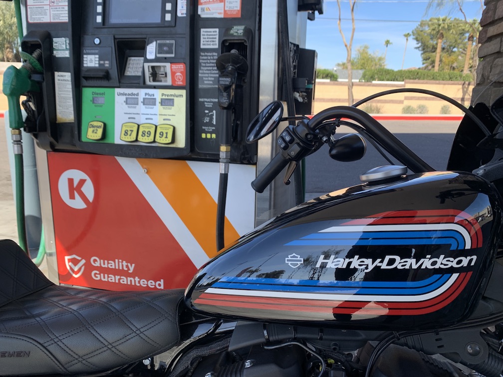 In the foreground the gas tank and seat of a black Harley-Davidson motorcycle, in the background a gas pump at a Cicle K, with two hoses, one green and one black, plus buttons to select the desired fuel and LCD displays for price and for purchasing fuel