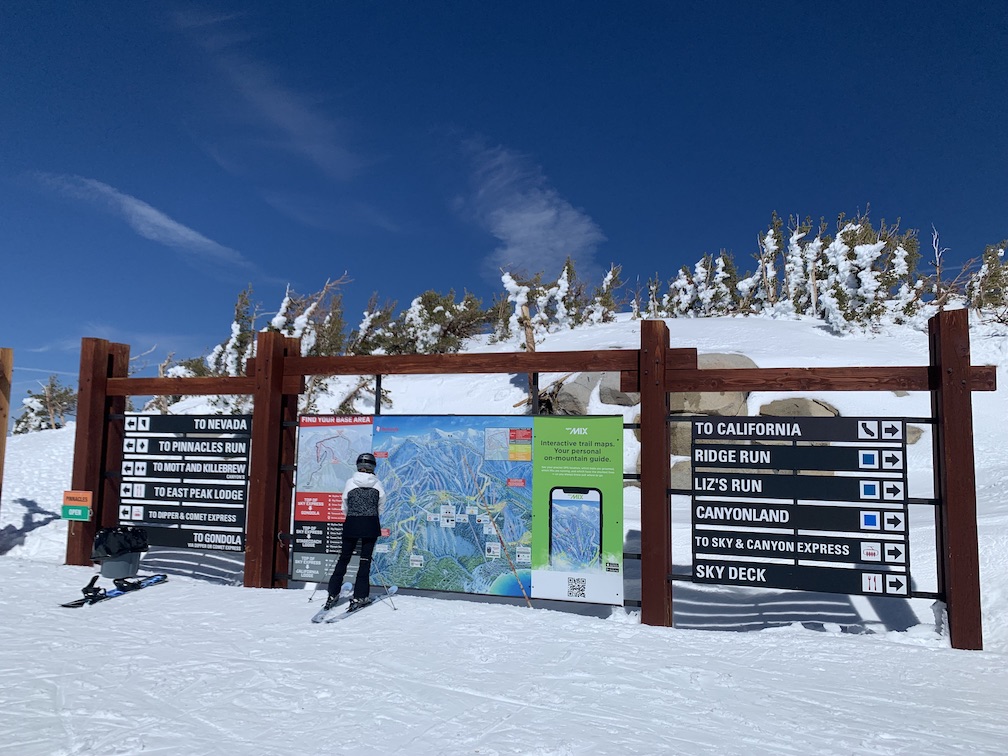 A skier standing in front of a trail map just below the peak of a ridge, with a set of sign planks to the right of the map with the topmost saying 'to California' and a set of sign planks to the left of the map with the topmost saying 'to Nevada'