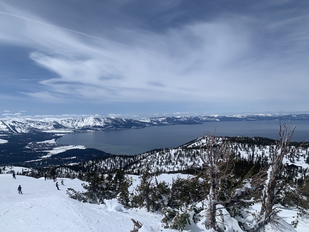 In the background a large mountain lake surrounded by snow-covered mountains, in the foreground a wide ski slope with a few skiers and snowboarders