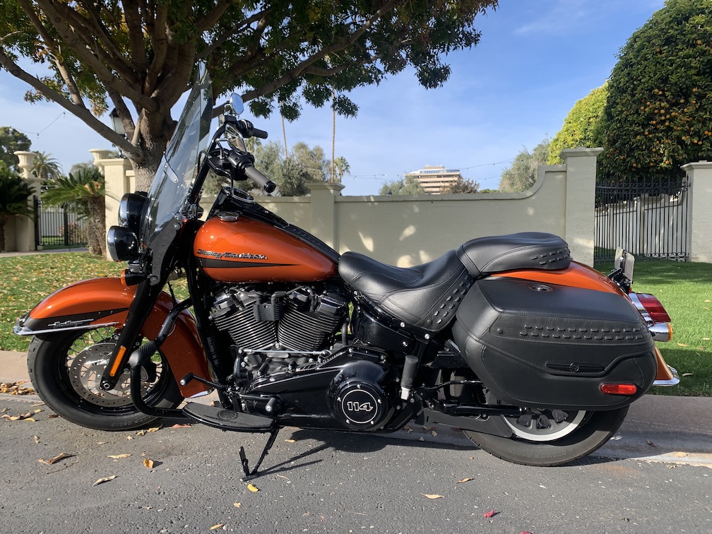 An orange and black 2020 Harley-Davidson Heritage Classic 114 motorcycle, viewed from the left side while parked on a sunny street