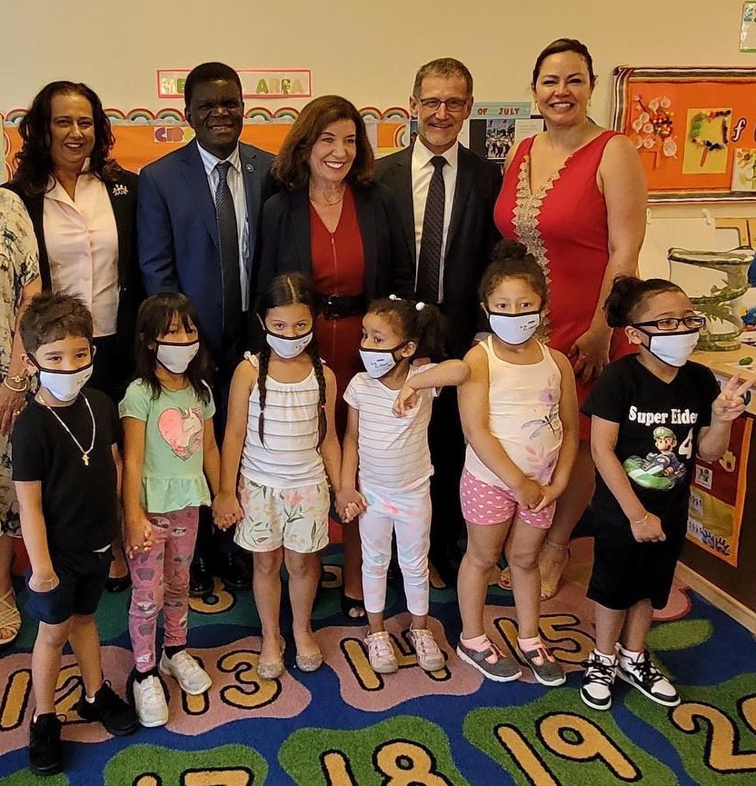 A row of six very young schoolkids wearning masks standing in a school classroom, with a row of five smiling, unmasked adults behind them