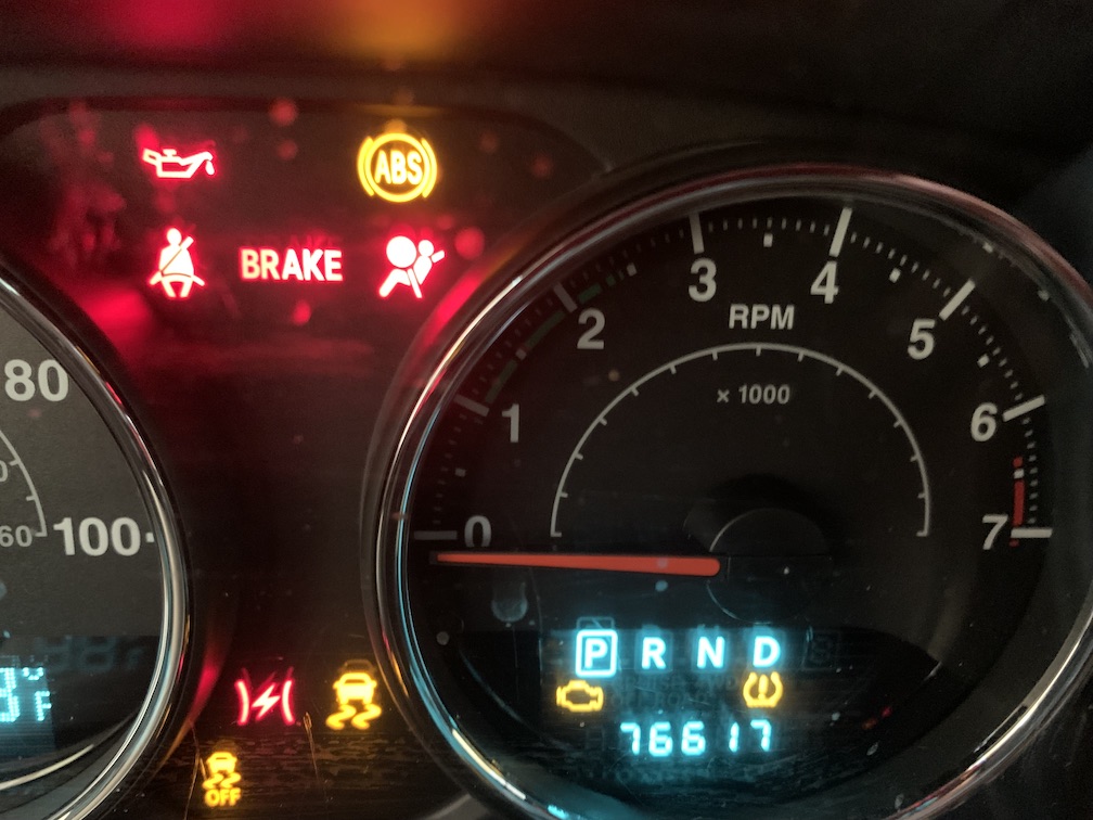 A portion of a black vehicular dashboard, with most of a circular RPM guage visible on the right, with an odometer and gear indicator inside, along with ten different icon lights lit up in either red or yellow to the left of the guage or inside of it