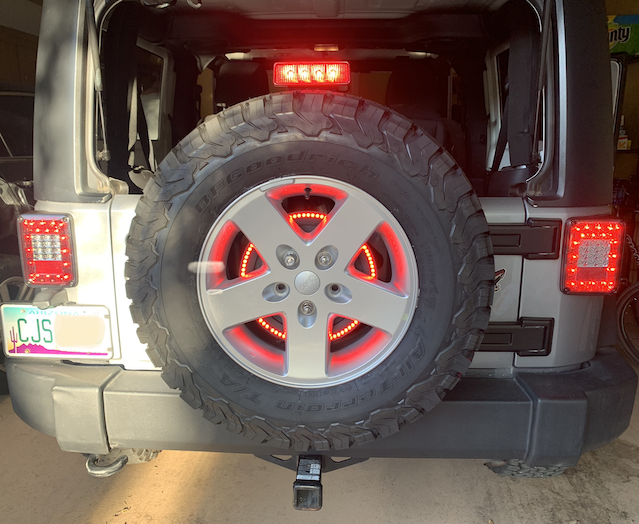 The back end of a grey Jeep Wrangler with four rear indicator lights illuminated: driver's side and passenger's side LED lights, a light above the exposed spare tire, and a ring of LED lights behind the spare tire and visible through gaps in the wheel