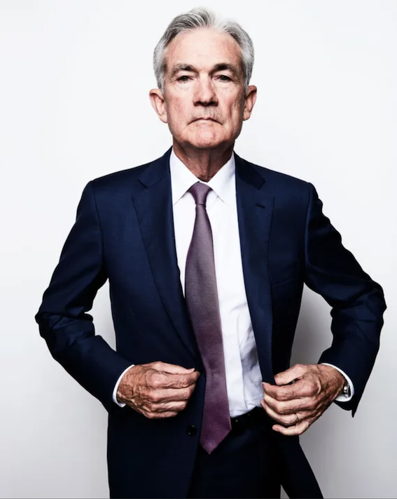 A grey-haired man adjusting the jacket of his blue suit, with white shirt and lavender tie