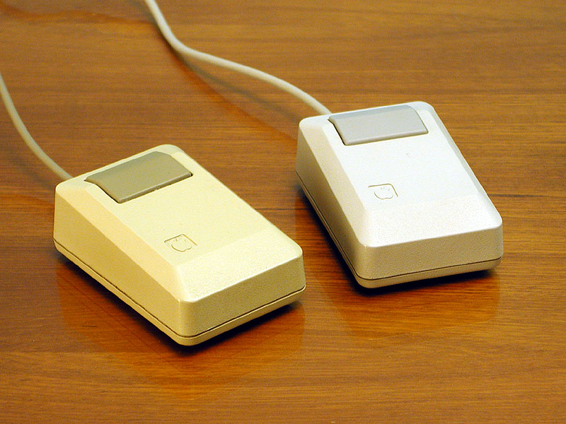 Two beige, angular, block-shaped computer mice, each with a single light brown button and a cord, pictured while resting on a wooden surface