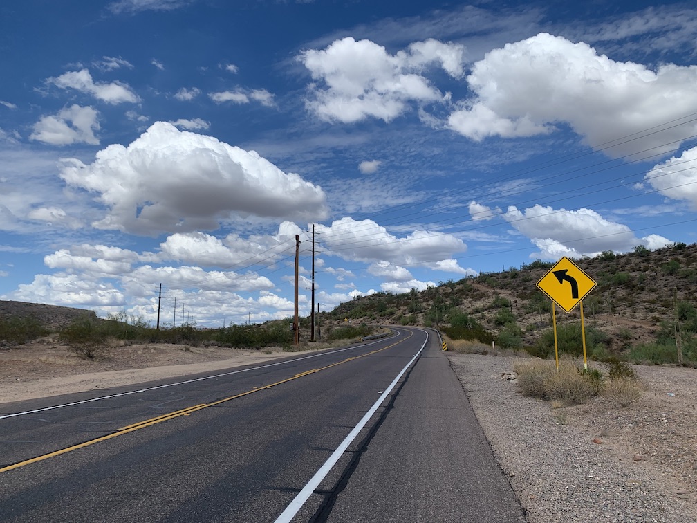 A two lane asphalt road that curves to the left before it reaches a hill in a desert landscape, with a yellow road sign indicating the curve to the left