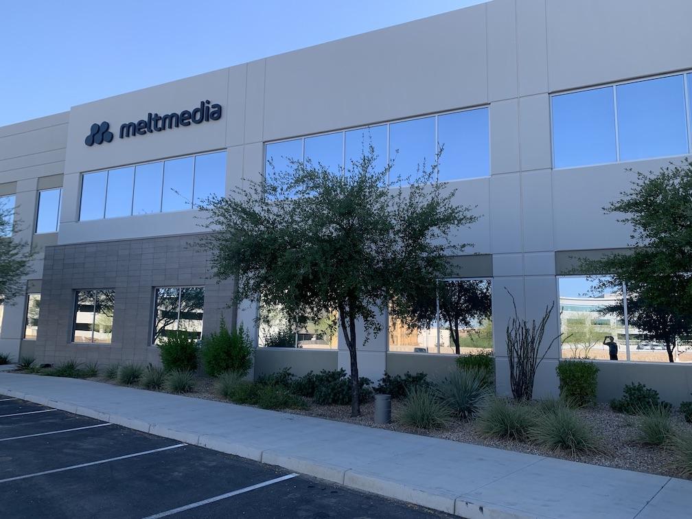 A portion of the exterior of a modern two-story office building, with a company logo and name afixed to the second story exterior wall, with reflective glass on the large windows, and in the foreground some plantings and parking spaces