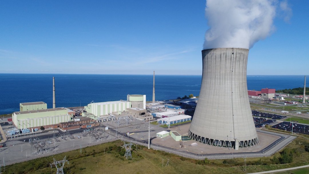 In the foreground an industrial plant consisting of a concrete tapered cylinder hundreds of feet tall emitting steam from the top, with other mostly windowless buildings nearby along with an asphalt parking lot with some cars, and in the background a huge expanse of blue water with nothing else visible on the horizon, on a sunny day with blue skies