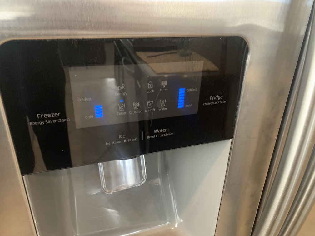 An LED refrigerator control panel which allows the freezer and chiller to be set between 'cold' and 'coldest', with a few other LEDs for other features, below which is a dispenser for ice and water