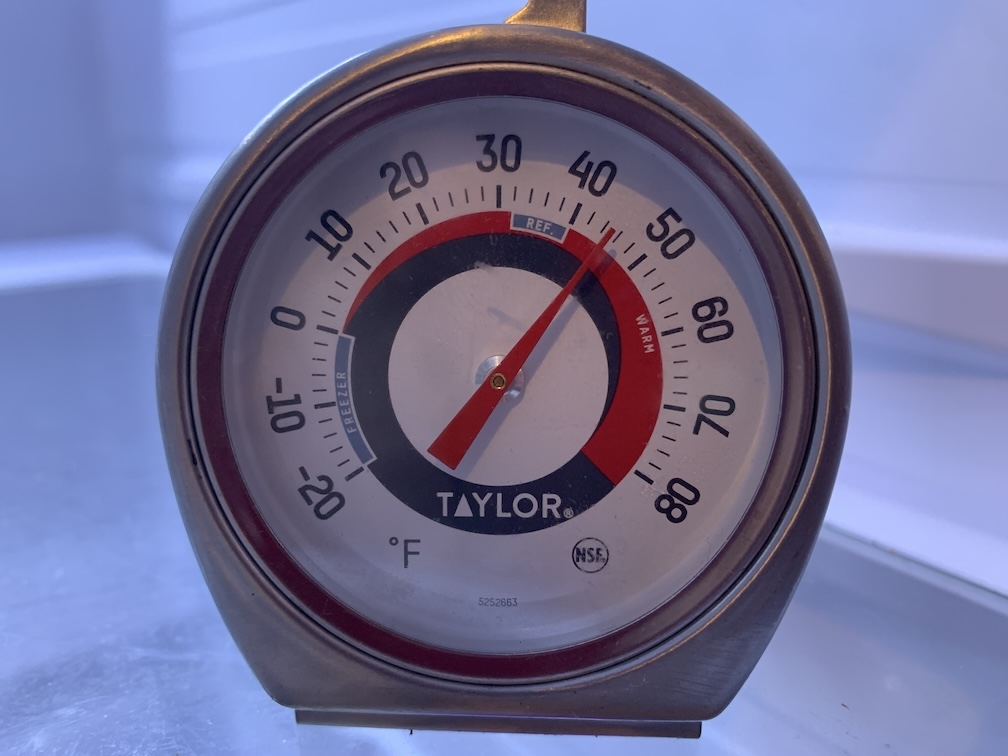 A round thermometer dial in a metal frame, inside a white compartment, with a red needle pointing midway between 40 and 50 degrees Fahrenheit, in the zone labeled 'warm'