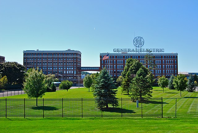 In the foreground, some city blocks with nothing on them but green grass, trees, and fencing; in the background, two approximately 7-story brick buildings with lots of windows, one of which has a large sign with the words 'General Electric' and the company's logo on top of it