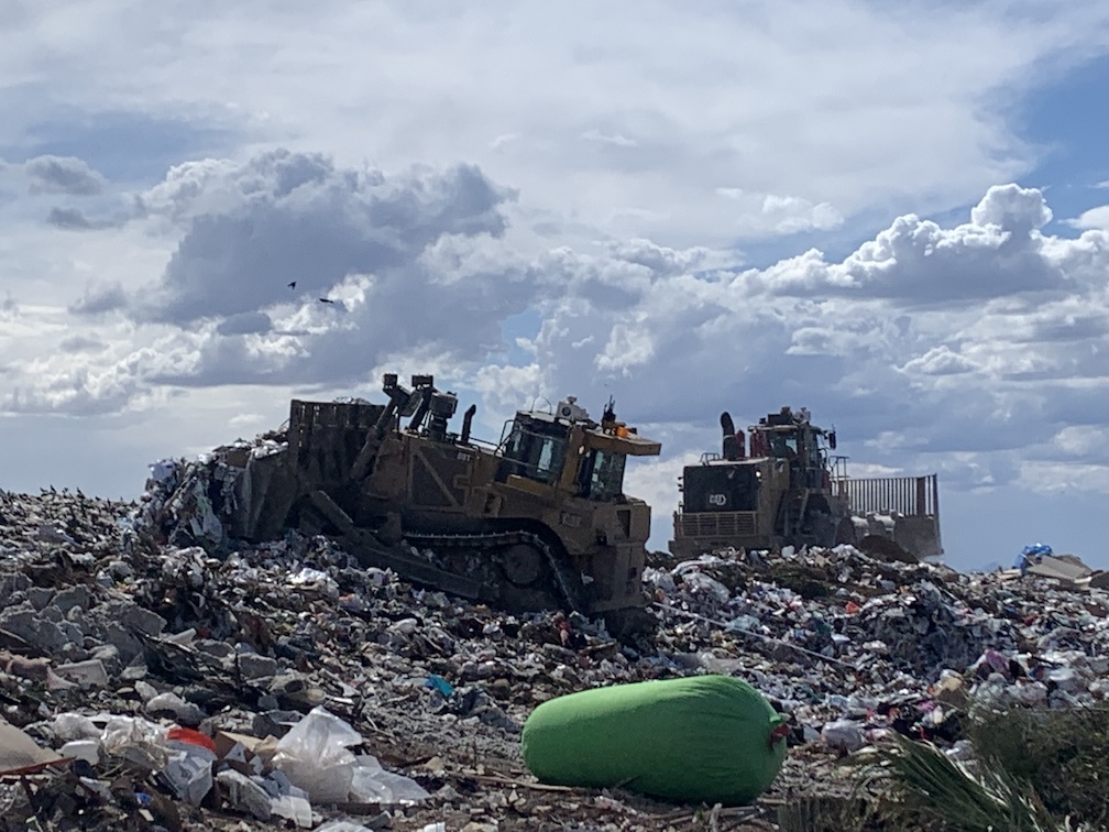A large heap of trash upon which two yellow bulldozers are rearranging trash while a few birds circle overhead on a partly sunny day