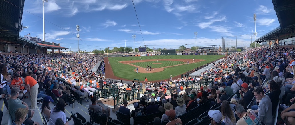 A panoramic view of a single-tiered baseball stadium taken from behind home plate during a baseball game, with nearly all the seats filled with fans