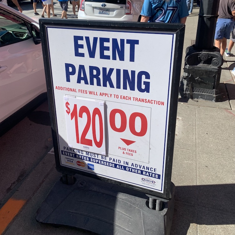A sidewalk-level sign for event parking, with a price of $120 in bold red numbers, and people walking on the sidewalk and street in the background