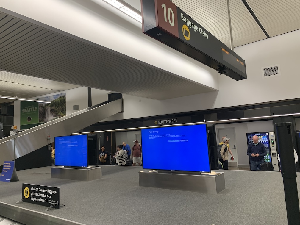 A closeup of an airport baggage claim, with a metal chute in the background and two dsplay screens in the foreground, both of which are bright blue with some text on them with the word 'recovery' being the largest