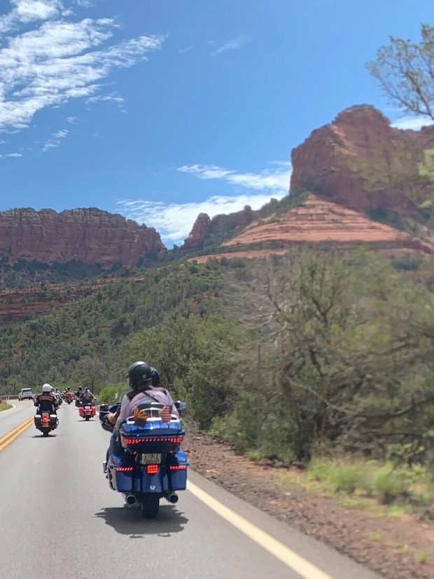 A group of motorcyclists riding on a road that turns to the left, alongside red sandstone cliffs and hills, partially covered with vegatation