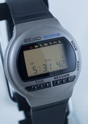 A gray-faced digital watch with a black band, displaying the time-of-day on the monochrome liquid crystal display, with several black buttons and labels on the face and side