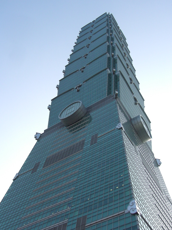 A view of a tall modern skyscraper broken up into multiple sections and clad in light green glass, viewed while looking up at the building from one corner at the base