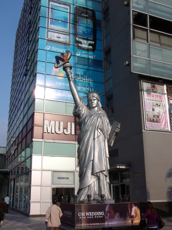An approximately 30 foot tall, gray version of the Statue of Liberty on a pedestal on a city street corner, with a modern glass building in the background, with advertising for consumer products displayed in some of the windows