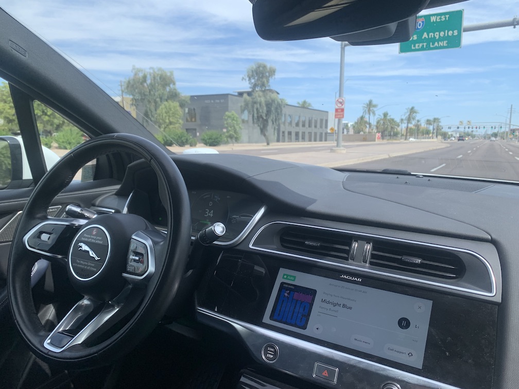 A picture taken from inside a car, showing the driver's wheel with no hands on it and the driver's side of the dashboard, while through the windshield is visible a three lane divided road that the car is driving down the middle of, with a sign for the entrance to an interstate highway overhead