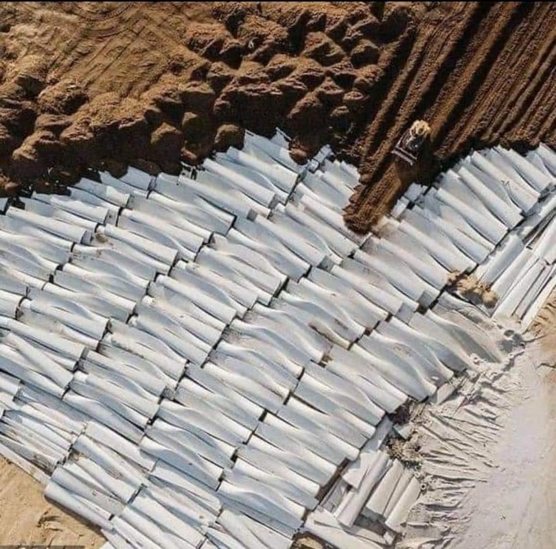 A picture taken from several hundred feet above showing rows of white windmill blade sections lying next to each other, hundreds in all, while in the upper portion a bulldozer is starting to push dirt over the blade sections